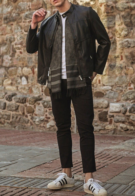 Person wearing a black vintage leather jacket, scarf, and Adidas sneakers against a stone wall.