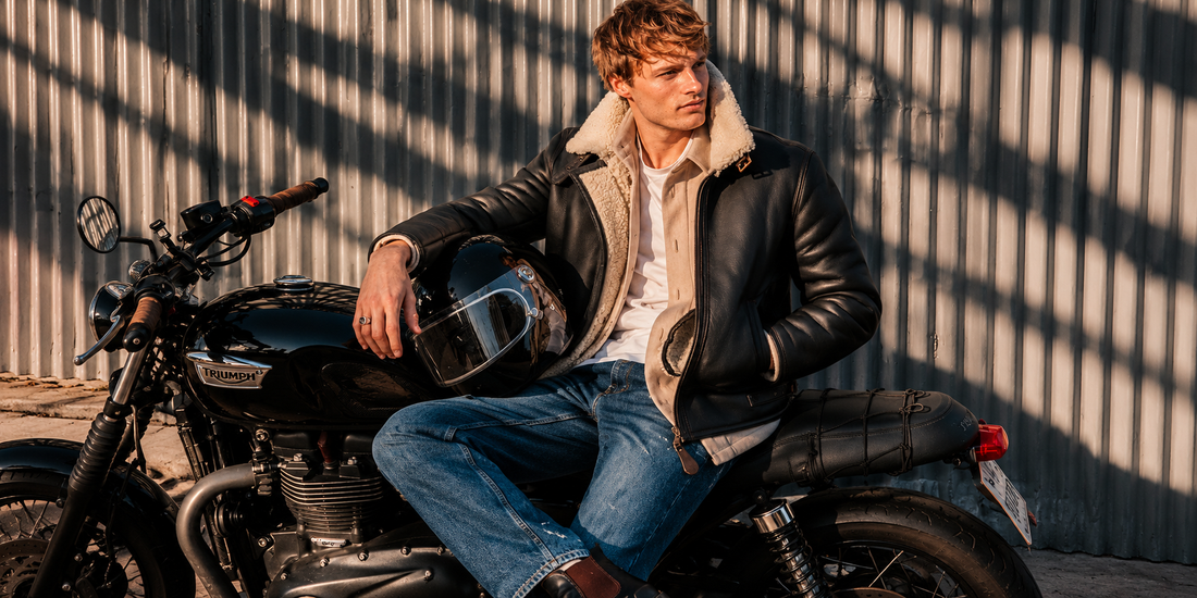 Man wearing a black shearling-lined leather aviator jacket and blue jeans, sitting on a motorcycle with a helmet in an urban outdoor setting.