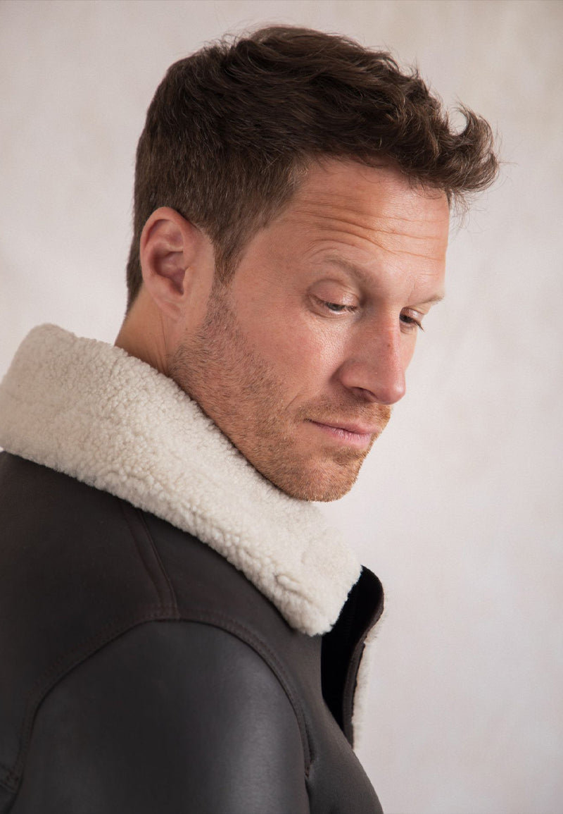 A man wearing a men's shearling leather jacket with a white fur collar against a plain background