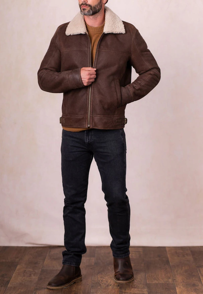Man wearing a brown leather jacket with a shearling collar and dark jeans against a plain background