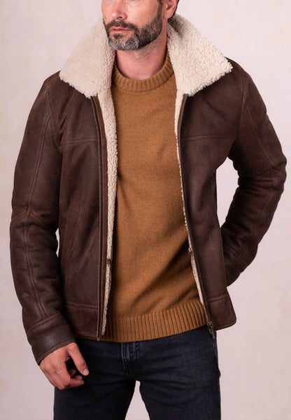 Man wearing a brown shearling leather jacket with a white fur collar and a brown sweater against a plain background