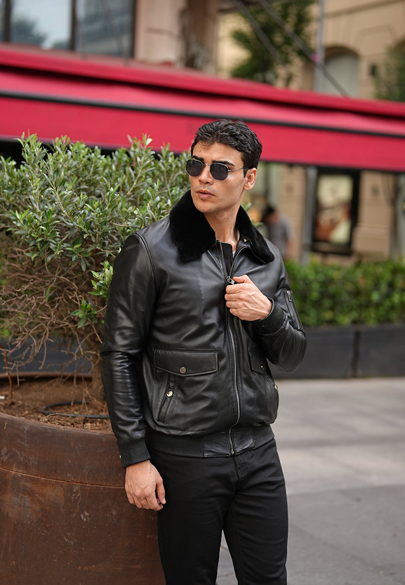 A man wearing a black Sheepskin leather jacket and sunglasses is standing outdoors with a red building in the background.