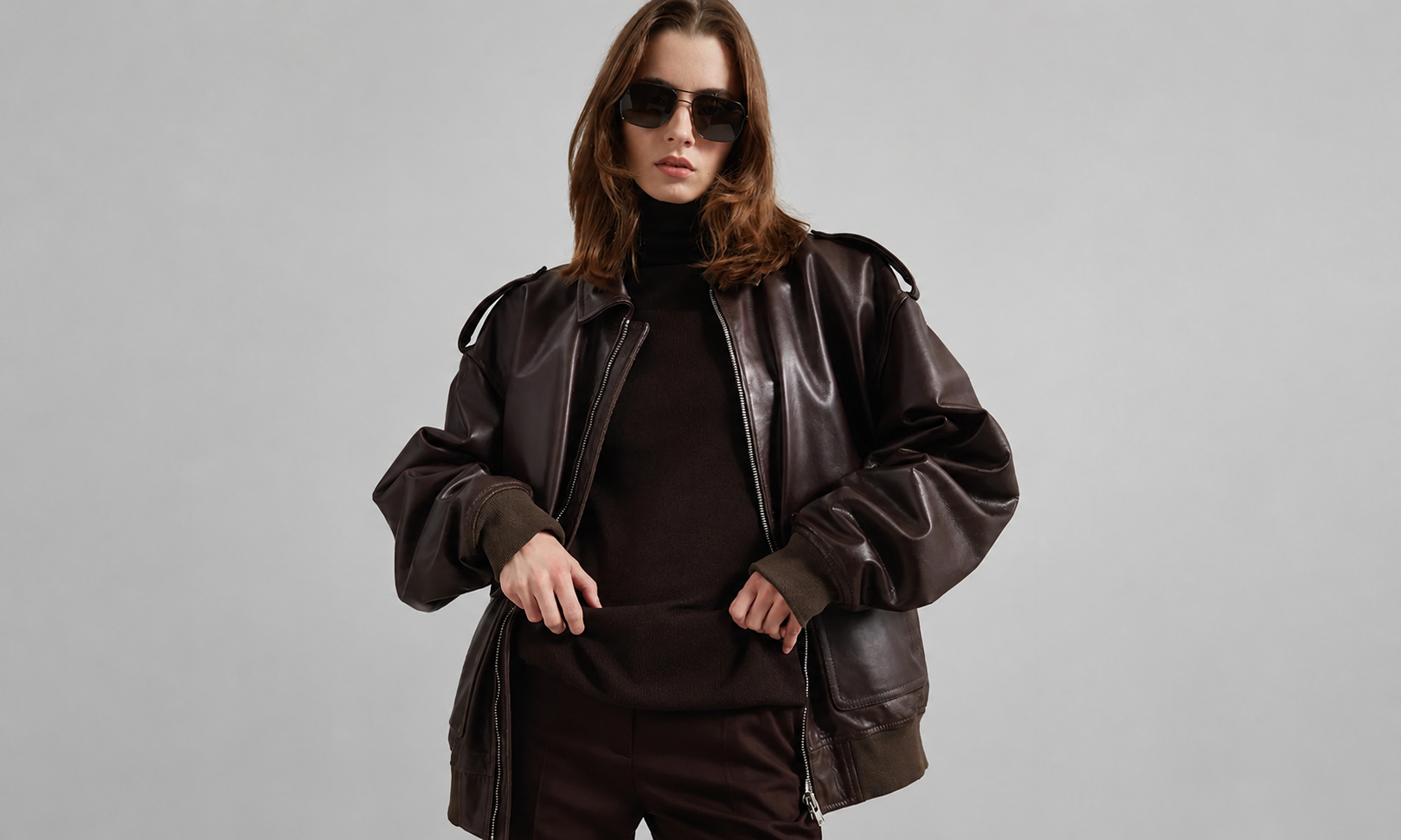 Woman wearing an oversized dark brown leather bomber jacket and black turtleneck, posing in a studio against a light gray background while wearing sunglasses.