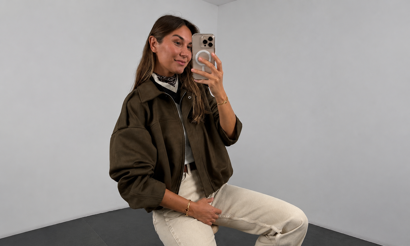 Woman taking a mirror selfie wearing a brown sheepskin leather bomber jacket and white pants against a plain background